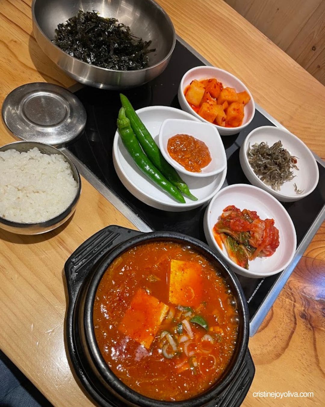 Bubbling red kimchi jjigae with tofu in a black pot, served with rice and Korean side dishes