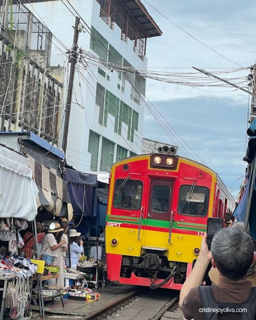Red and yellow SRT trains rolling through market stalls at Maeklong Railway Market as visitors film it