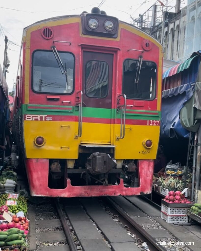 Red and yellow SRT train 1227 approaching on the tracks at Maeklong Railway Market, with fruit stalls on both sides