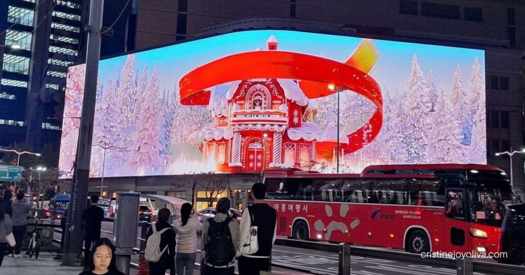 3D digital billboard of a gingerbread house with snow-covered trees in Myeongdong at night