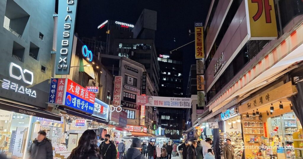 Shoppers on a Namdaemun Market street at night, lined with neon signs, eyeglass shops, and dried-food stalls