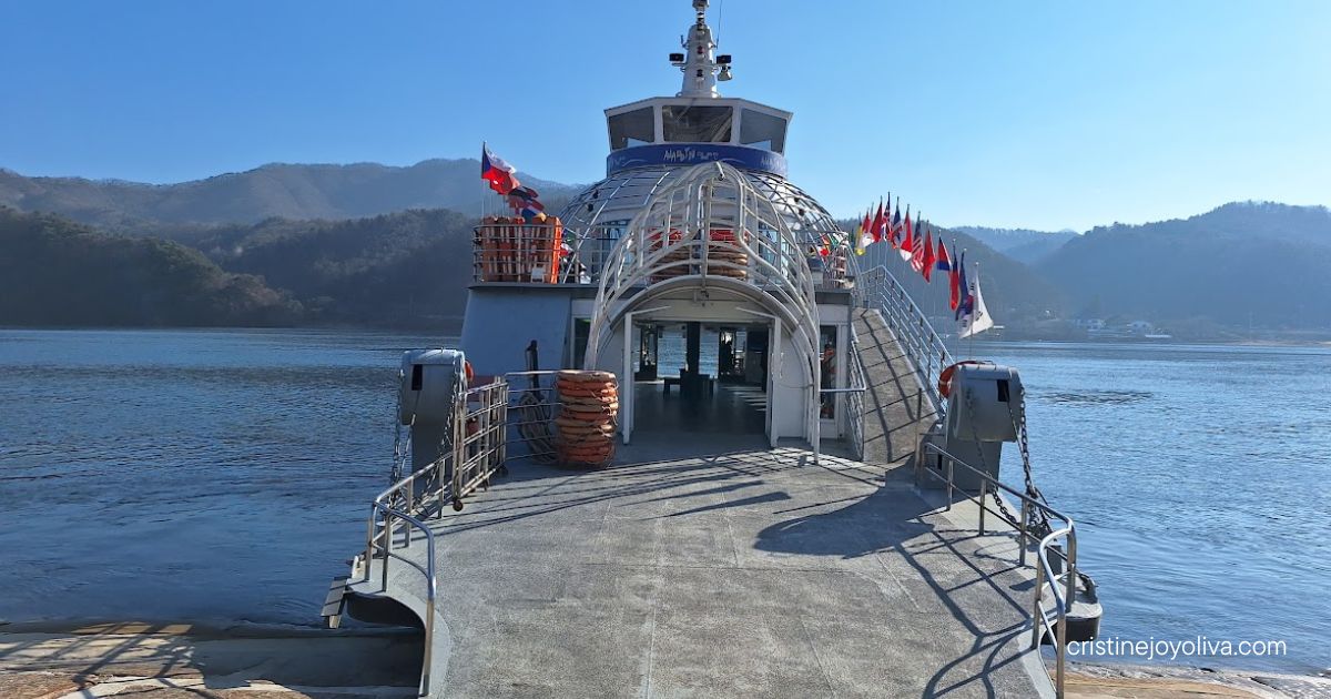 Ferry docked at a stone pier on a lake with international flags, life rings, and forested mountains under a clear blue sky