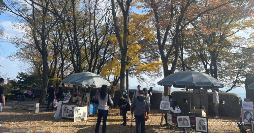 Portrait artists with umbrellas and framed drawings under yellow autumn trees in Namsan Park, Seoul