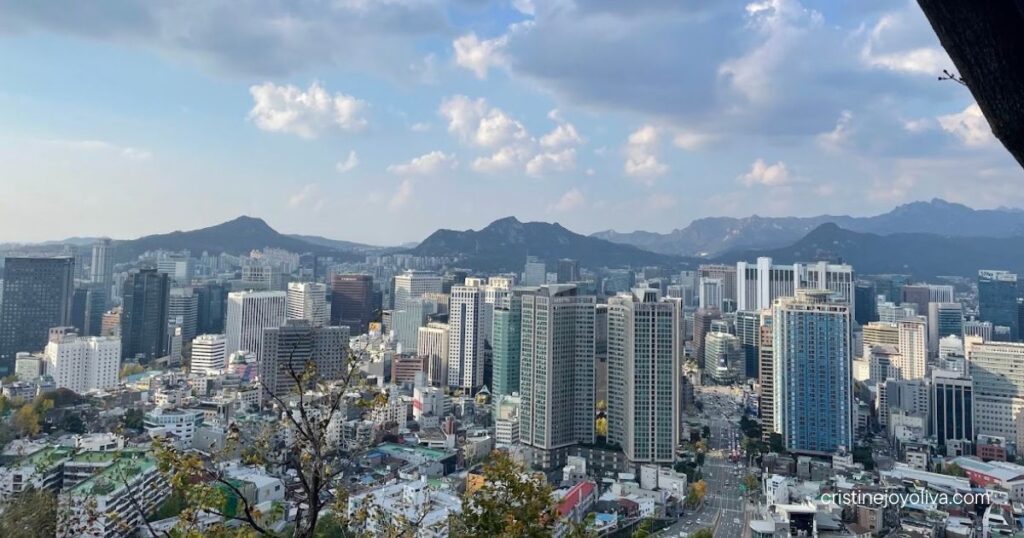 Seoul skyline with high-rises and distant mountains seen from Namsan under a partly cloudy sky