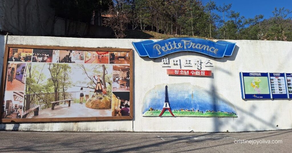 Petite France sign with Eiffel Tower mural, photo panel, and Gapyeong tourist map on a white wall under a blue sky