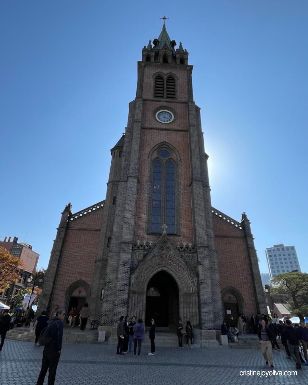 Myeongdong Cathedral's red brick clock tower and arched entrance under a blue sky, with visitors outside