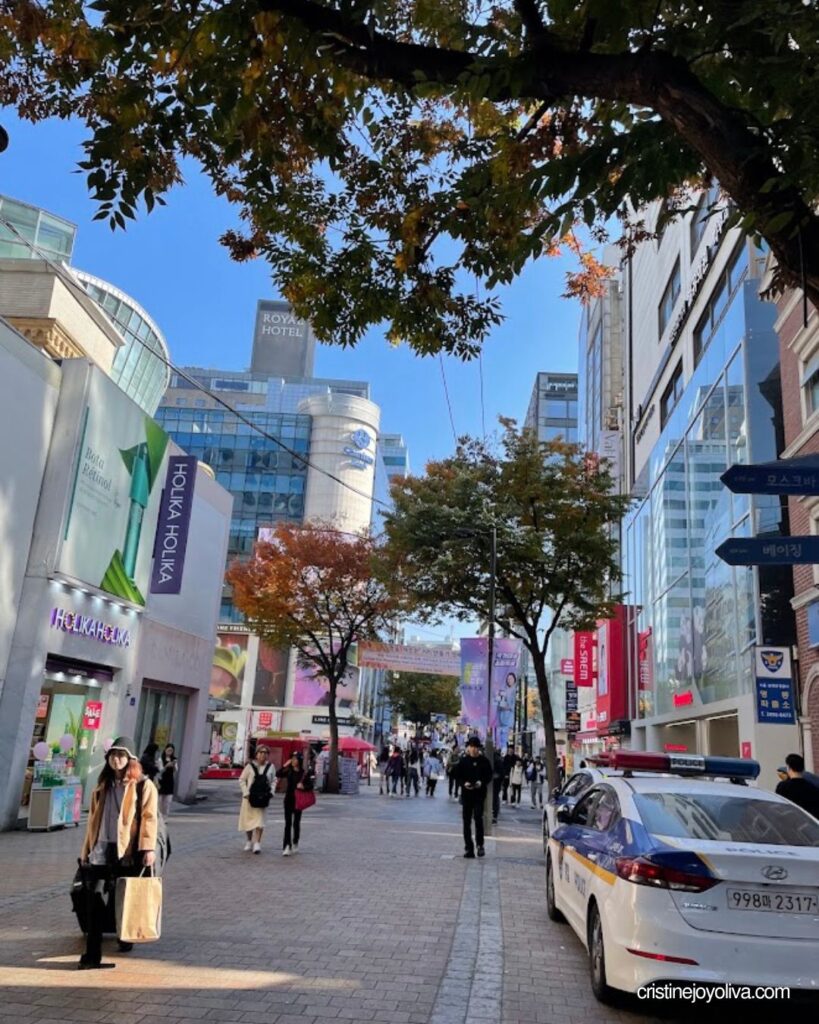Shoppers walking along Myeongdong Street in Seoul under autumn trees, with storefronts and a police car nearby