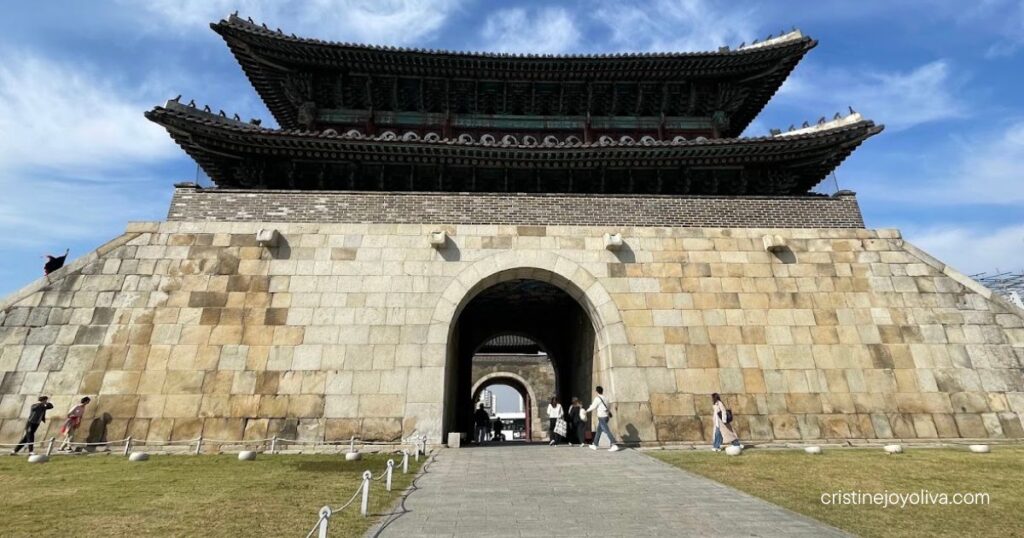 Hwaseong Fortress gate with an arched stone entrance and two-tiered wooden pavilion under a blue sky, visitors walking through
