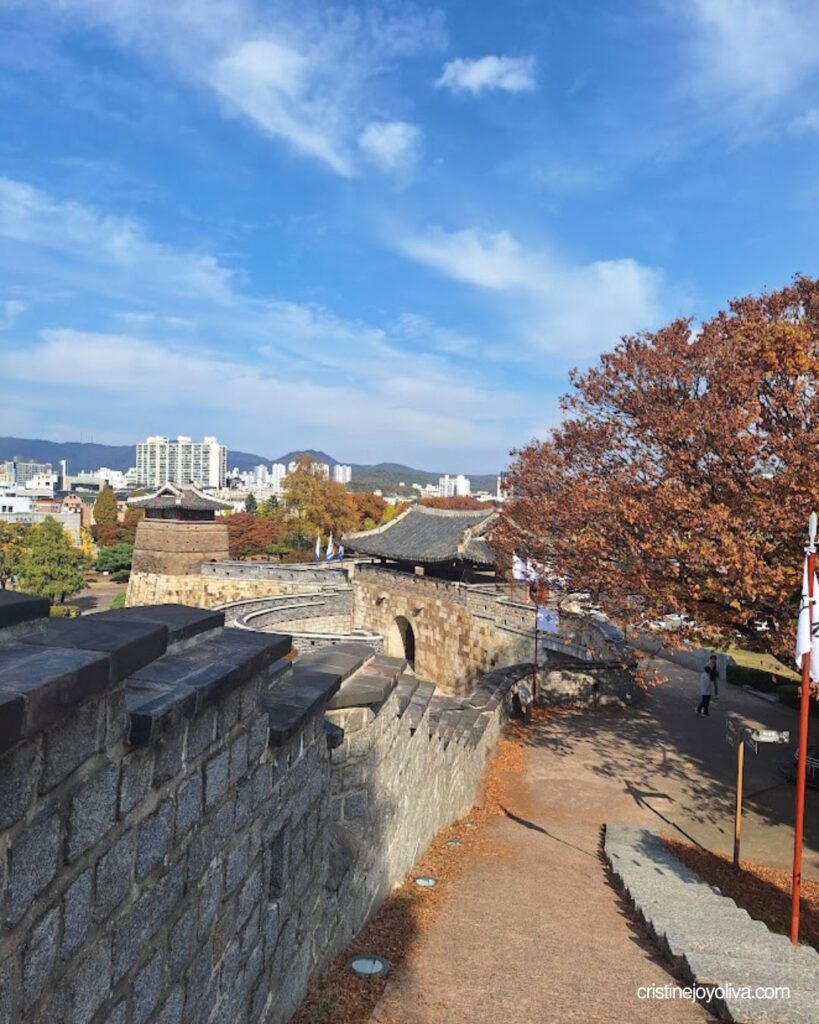Curving stone wall of Suwon Hwaseong Fortress with arched gateways, tiled rooftops, and autumn trees above the city