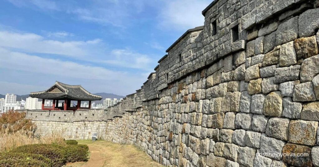 Stone wall of Suwon Hwaseong Fortress beside a dirt path with tall dry grass and a tiled-roof pavilion under a blue sky