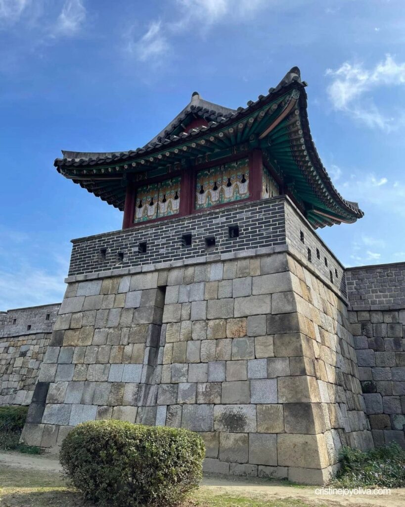 Painted wooden watchtower with a curved tiled roof on a stone and brick base of Suwon Hwaseong Fortress under a blue sky