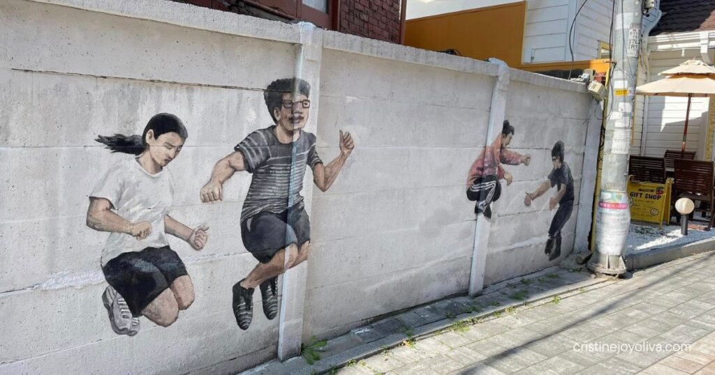 Street mural of four people jumping mid-air on a white wall along a paved alley beside a gift shop sign