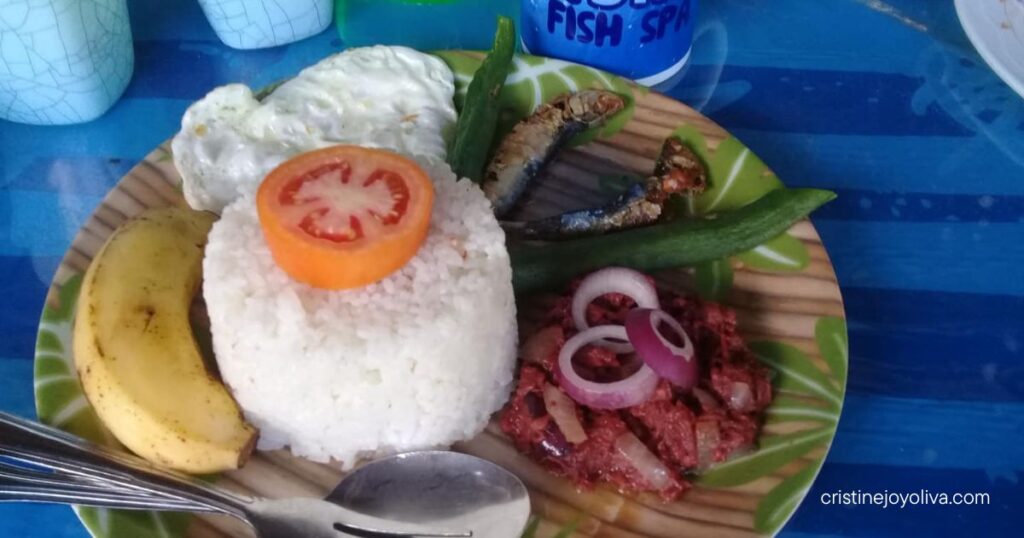 Plate of traditional Filipino breakfast in Antique, Philippines, with rice, fried egg, small fried fish, corned beef with onions, okra, tomato, and banana