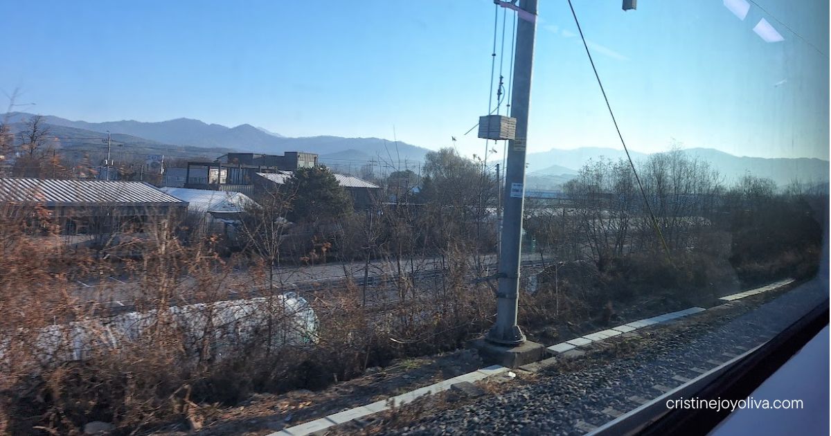 View from a train window of railway tracks, farm buildings, bare trees, and mountains under a clear blue sky in winter